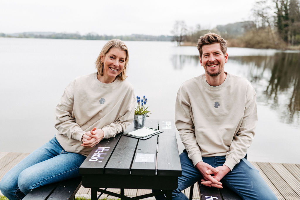 Zwei Personen sitzen an einem Tisch am Ufer eines Sees, beide tragen beige Sweatshirts.