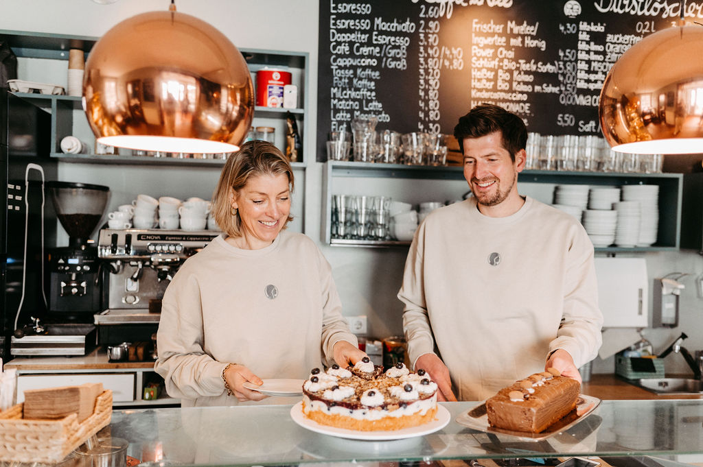 Zwei Mitarbeiter einer Konditorei präsentieren Kuchen in einem Café mit einer modernen Einrichtung.